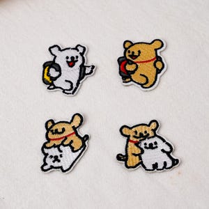 Op de afbeelding: Vier geborduurde patches met tekenfilmfiguren. Een witte beer met een gele tas, een beige beer met een rode halsband, een beige beer die op een witte beer zit en een beige beer die een witte beer knuffelt. Elke patch heeft een witte rand.