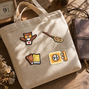 May include: A beige canvas tote bag featuring four embroidered patches: a brown owl holding a letter, a wooden broomstick, two books, and a yellow sign with the text 9 3/4. The bag has two fabric handles and a simple, rectangular construction.