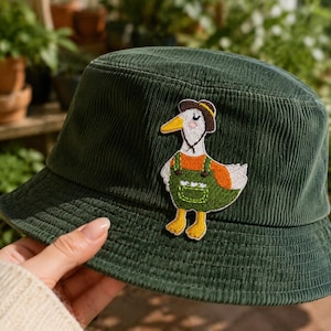 May include: A dark green corduroy bucket hat featuring an embroidered patch of a white duck wearing a brown hat and green overalls. The duck has an orange wing, yellow feet, and a small pocket detail on the overalls, centered on the front panel.