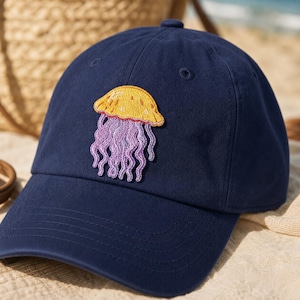 May include: A navy blue baseball cap featuring an embroidered jellyfish design on the front. The jellyfish has a yellow bell with small white dots and long, wavy purple tentacles. The cap has a curved brim and stitched eyelet details.