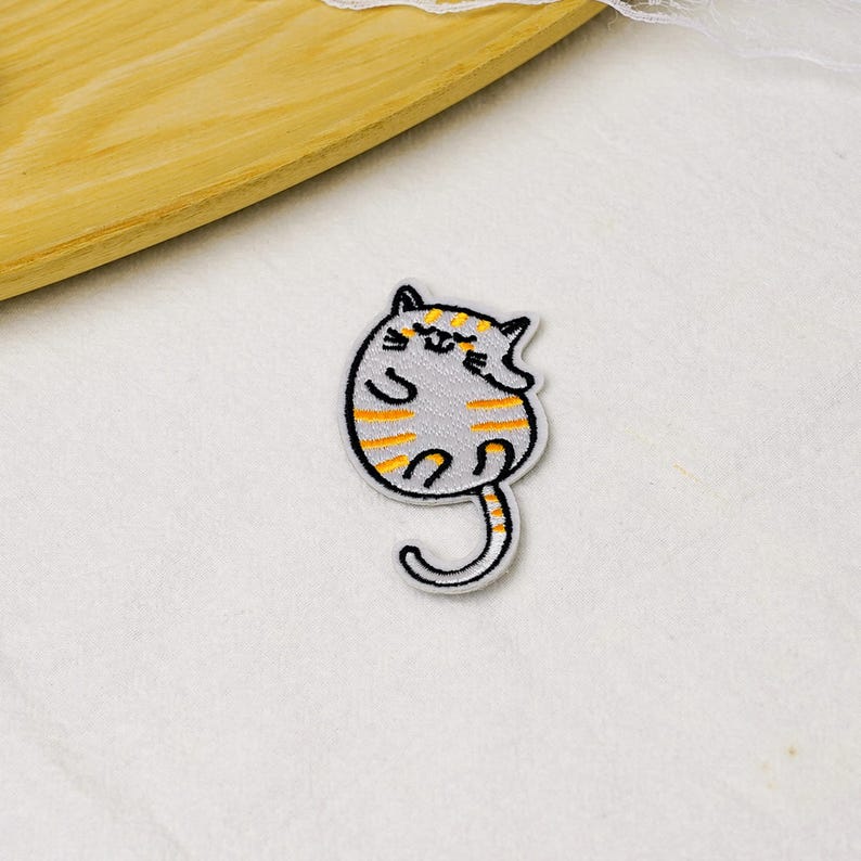 May include: An embroidered patch featuring a round, light gray cat with orange stripes, a long curved tail, and a simple black outline. The cat has closed eyes and a small smile, depicted in a curled, resting position with its paws tucked in.