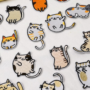 May include: A collection of embroidered cat patches featuring various feline designs in colors including orange, gray, brown, and white. Each patch depicts a cartoon-style cat in different poses, such as sitting, lying down, or stretching, with stitched details.