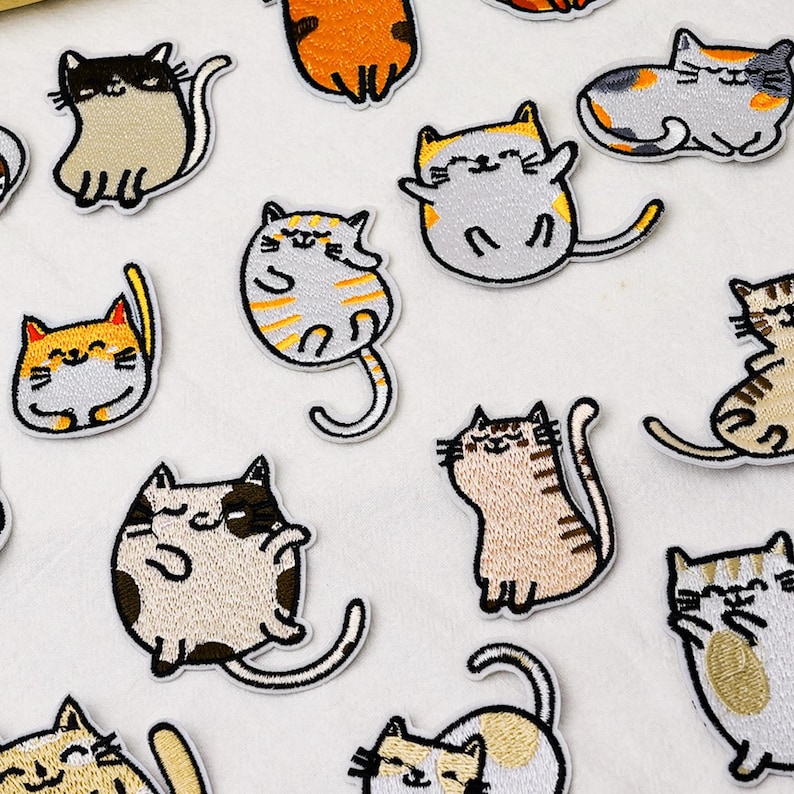 May include: A collection of embroidered cat patches featuring various feline designs in colors including orange, gray, brown, and white. Each patch depicts a cartoon-style cat in different poses, such as sitting, lying down, or stretching, with stitched details.