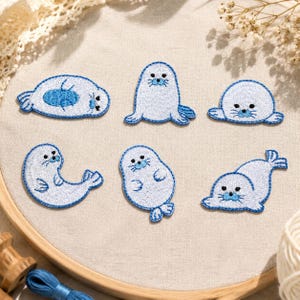 May include: Six embroidered patches featuring white and light blue seals in various poses. Each seal has black eyes, a blue nose, and stitched whiskers. The patches are arranged in two rows of three, showcasing different orientations and movements.