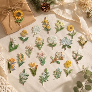 May include: A collection of various embroidered flower patches featuring diverse botanical designs in shades of yellow, white, blue, and green. Each patch depicts a different floral species with detailed stitching, arranged on a light fabric surface.