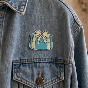 May include: An embroidered patch shaped like a round gift box with a light blue base and a cream-colored ribbon tied in a bow on top. The patch is attached to the chest area of a light blue denim jacket featuring a metal button on the pocket.
