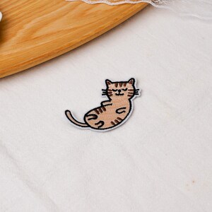 May include: An embroidered iron-on patch featuring a light brown tabby cat with dark brown stripes, lying on its back with eyes closed and a small smile. The cat has a white border outline and is depicted in a relaxed, curled position.