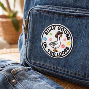 Peut inclure: Un sac à dos en jean bleu avec un écusson brodé circulaire. L'écusson représente une oie blanche portant un chapeau de cowboy marron, entourée de petites fleurs colorées et du texte noir "SILLY GOOSE ON THE LOOSE" en bordure circulaire.