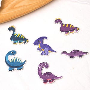 Puede incluir: Un conjunto de seis parches bordados de dinosaurios en varios colores y formas. El set incluye dos dinosaurios azules con rayas amarillas, dos dinosaurios morados con manchas amarillas o oscuras, un dinosaurio bípedo y un dinosaurio de cuello largo.
