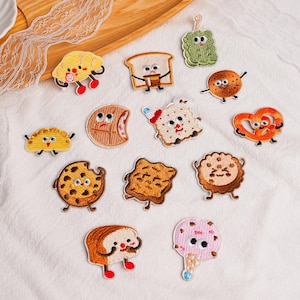 Könnte beinhalten: Eine Sammlung von zwölf bestickten Aufnähern mit anthropomorphen Lebensmitteln, darunter ein Croissant, Toast, eine Gewürzgurke, Kekse, eine Brezel, ein Teigtäschchen und eine Eistüte, jeweils mit Cartoon-Augen und Gliedmaßen auf weißem Stoff.