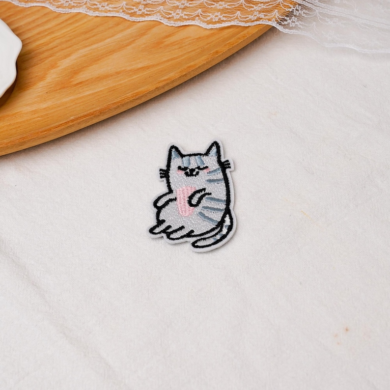 May include: A gray and white embroidered cat patch featuring a seated feline with dark gray stripes, closed eyes, and a small pink heart shape on its chest. The patch has a black stitched outline and a white border, showcasing detailed thread work.