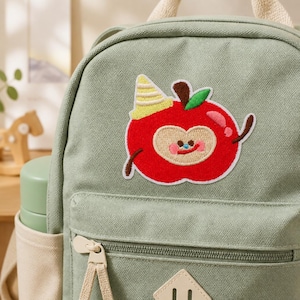 May include: A sage green canvas backpack featuring an embroidered patch of a red apple character wearing a yellow party hat. The apple has a tan face, a small smile, and a green leaf on top. The backpack includes a front zippered pocket with beige pulls.