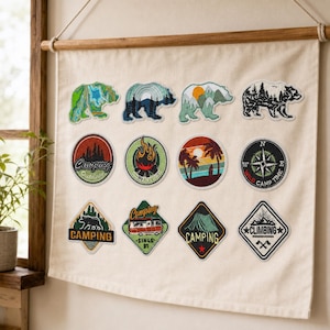 May include: A cream-coloured fabric wall hanging displaying twelve embroidered patches with outdoor themes. The patches feature designs of bears, a campfire, a compass, a camper van, a tent, and mountains, with text including Camping and Climbing.