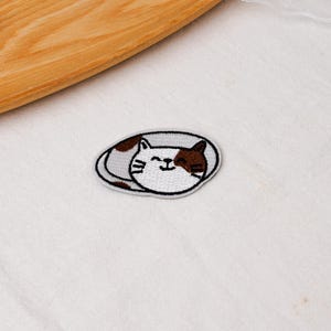 May include: An oval-shaped embroidered patch featuring a white and brown cat curled into a sleeping position. The cat has closed eyes, a small smile, and visible whiskers, with brown patches on its head and body, all outlined in a dark grey border.
