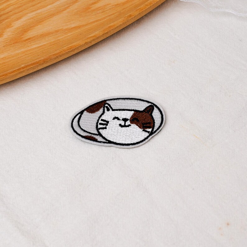 May include: An oval-shaped embroidered patch featuring a white and brown cat curled into a sleeping position. The cat has closed eyes, a small smile, and visible whiskers, with brown patches on its head and body, all outlined in a dark grey border.