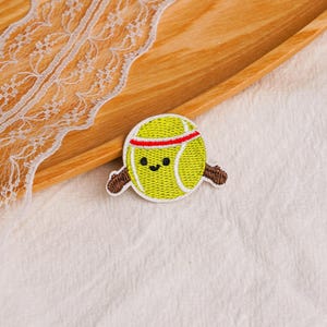 May include: A circular embroidered patch featuring a yellow tennis ball with a smiling face, two small black eyes, and a red horizontal stripe across the top. The tennis ball is flanked by two brown wooden-textured baseball bats on each side.