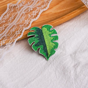 May include: A monstera leaf patch featuring detailed embroidery in shades of dark forest green and vibrant lime green. The leaf has a distinct split-leaf design with serrated edges and a white border, showcasing a textured, stitched surface.