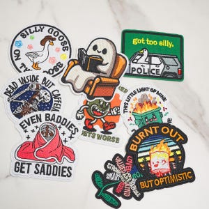 Op de afbeelding: Een verzameling geborduurde patches met diverse ontwerpen, waaronder een gans, een lezend spook, een politieauto met een gans, een skelet, een brandende afvalcontainer, een persoon in een deken en motiverende teksten.