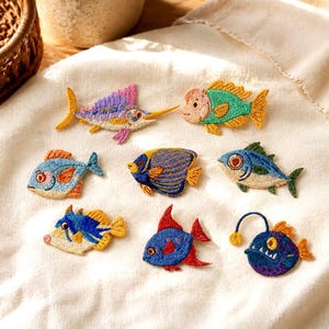 May include: A collection of eight colourful embroidered fish patches featuring various species including a swordfish, an angelfish, and an anglerfish. Each patch displays intricate thread work in vibrant shades of blue, yellow, red, and purple.