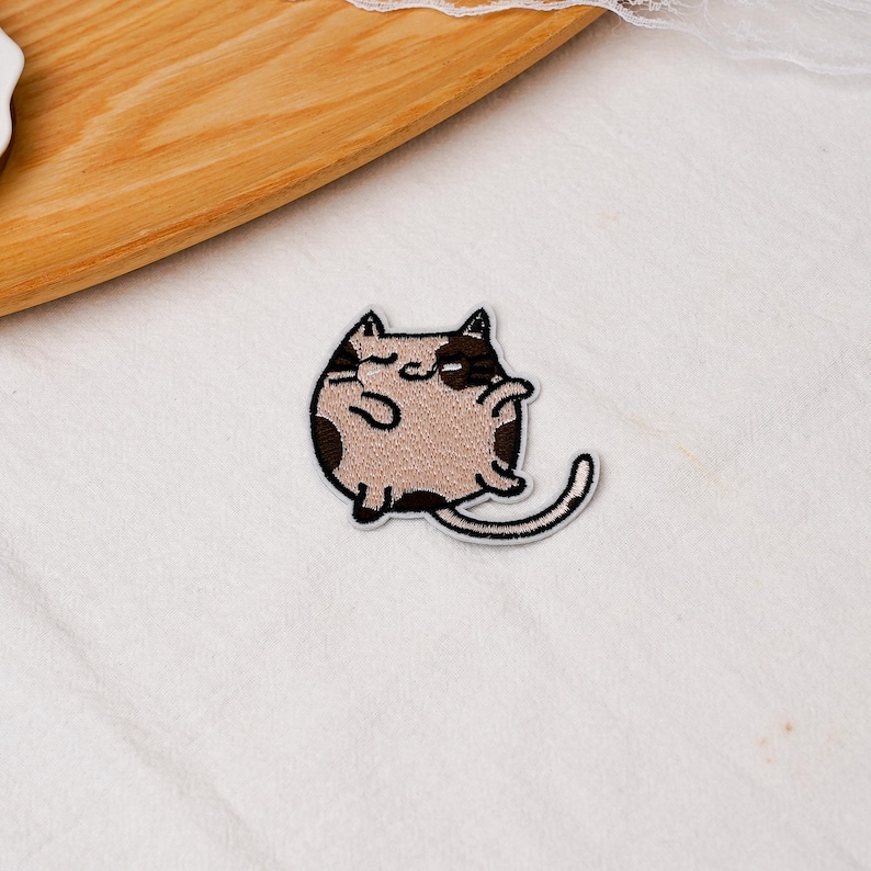 May include: An embroidered patch featuring a round, beige cat with dark brown patches on its head, back, and tail. The cat has a simple black outline, small ears, and a long, curved tail, rendered in a flat, cartoon-style design with stitching.