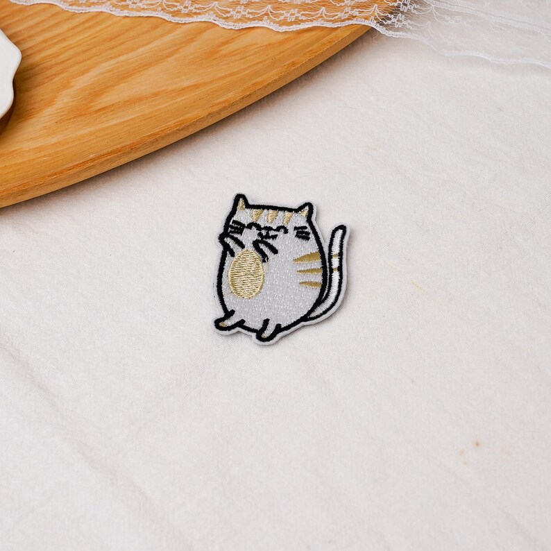 May include: An embroidered patch featuring a cartoon cat with a light gray body, gold-colored stripes, and a gold belly patch. The cat has a black stitched outline, small ears, whiskers, and a curved tail, presented in a simple, flat design.