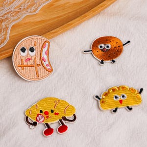 May include: Four embroidered fabric patches featuring cartoon food characters with faces. One is a tan circular cracker, one is a brown speckled cookie, one is a yellow crescent pastry, and one is a yellow folded dumpling with small limbs.