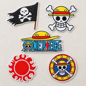 May include: Five embroidered patches featuring One Piece imagery: a black skull and crossbones flag, a skull wearing a yellow straw hat with crossbones, a blue One Piece logo, a red sun emblem, and a skull with a straw hat inside a yellow wheel.
