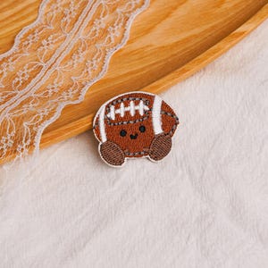 May include: A brown embroidered patch shaped like a football with white laces and stripes. The football features a cute, simple face with two small black circular eyes and a small curved black mouth, giving it a friendly and animated appearance.
