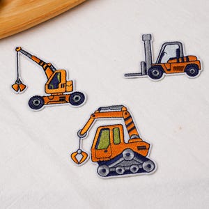 May include: Three embroidered patches featuring construction vehicles in orange, grey, and black. The set includes a crane with a hook, a forklift with a tall mast, and an excavator with a tracked base and a mechanical arm with a claw attachment.