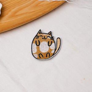 May include: An embroidered iron-on patch featuring a round, golden-brown cat with white spots. The cat has a simple black stitched outline, closed eyes, a small smile, and a long tail curved to the side, all set against a white fabric background.