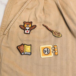 May include: Four embroidered patches featuring a brown owl holding a letter, a wooden broomstick, two books, and a yellow rectangular sign with the text 9 3/4. The patches are arranged on a tan fabric surface with visible diagonal stitching lines.