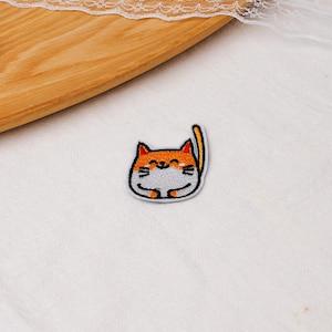 May include: An embroidered iron-on patch featuring a cartoon cat with an orange head, white face, and a small orange tail. The cat has closed eyes, a small smile, and black whiskers, all outlined in black thread on a white fabric background.