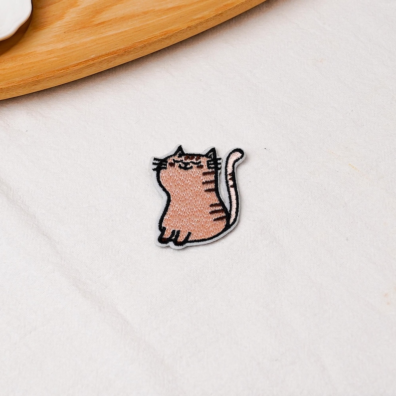 May include: A small embroidered patch featuring a light brown tabby cat with dark brown stripes, a curved tail, and a simple stitched face with closed eyes and a smile, outlined in black thread against a white fabric background.