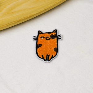 May include: An embroidered iron-on patch featuring a stylized orange cat with dark brown spots, black whiskers, and a simple black outline. The cat has a rounded body shape, small pointed ears, and a closed-eye expression with a small curved mouth.