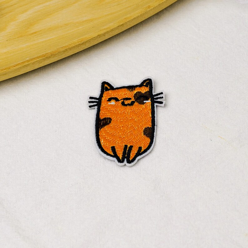 May include: An embroidered iron-on patch featuring a stylized orange cat with dark brown spots, black whiskers, and a simple black outline. The cat has a rounded body shape, small pointed ears, and a closed-eye expression with a small curved mouth.