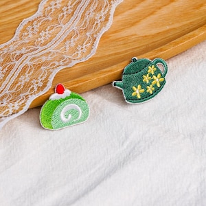 May include: Two embroidered patches featuring a green teapot with yellow floral details and a light green Swiss roll cake with a white cream center and a small red cherry on top, showcasing detailed stitching and vibrant thread colors.