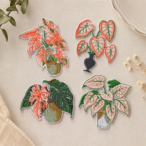 May include: Four embroidered fabric patches featuring potted houseplants. The designs include various leaf shapes in shades of pink, coral, and green, set in containers colored in olive green, black, and white, with detailed stitching throughout.