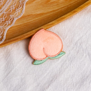 May include: A peach-shaped embroidered patch featuring a soft light orange colour with a darker orange outline. The design includes two small, textured green leaves at the base of the fruit, rendered with detailed stitching throughout the surface.