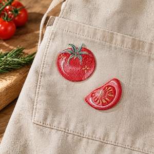 May include: A beige apron featuring two embroidered tomato designs on the front pocket. One design is a whole round red tomato with a green stem and white highlights, and the other is a red tomato wedge showing internal seeds and texture.