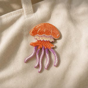 May include: An embroidered patch featuring a jellyfish design with a bright orange bell, a pale yellow and orange ruffled skirt, and long, slender tentacles in shades of purple and white, attached to a plain, light beige fabric surface.