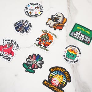 Puede incluir: Una colección de nueve parches bordados con diversos diseños, incluyendo un ganso, un fantasma leyendo un libro, un esqueleto, una bomba, un coche de policía, un contenedor de basura en llamas, una flor con texto, una persona en una manta y una nube de azúcar quemada.