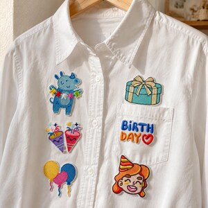 Puede incluir: Una camisa blanca de botones con parches bordados coloridos. Los diseños incluyen una criatura azul sosteniendo una pancarta, dos matasuegras, tres globos, una caja de regalo, el texto "BIRTH DAY" con un corazón y una persona con gorro de fiesta.