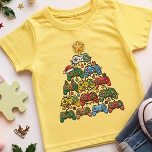 May include: A bright yellow short-sleeved t-shirt featuring a graphic print of a Christmas tree constructed from various colourful video game controllers, topped with a gold star, alongside blue denim jeans and blue canvas trainers.