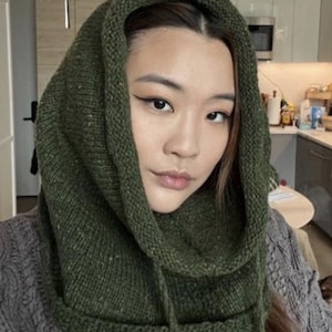 Könnte beinhalten: Eine grüne Strickhaube mit Kordelzug. Die Haube besteht aus einem strukturierten Garn und ist so konzipiert, dass sie Kopf und Hals bedeckt. Die Haube hat eine einfarbige grüne Farbe.