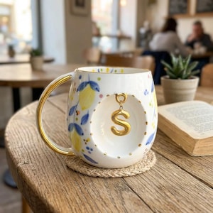 Può includere: Una tazza in ceramica bianca con un motivo dipinto di limoni gialli e foglie blu. La tazza presenta un manico color oro e un incavo circolare sulla parte anteriore contenente un ciondolo color oro a forma di lettera S appeso a un anellino.
