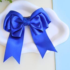 May include: A vibrant blue satin hair bow. The bow features a large, layered design with long, flowing tails. The center of the bow is embellished with a sparkling, textured material. This accessory is perfect for adding a touch of elegance to any hairstyle.