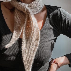 May include: A beige knitted scarf with a textured ribbed pattern tied in a knot around the neck. The scarf features a tapered design with pointed ends and a soft, fuzzy appearance, worn over a black long-sleeved ribbed top with a v-neckline.