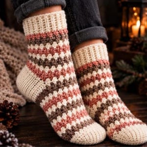 Könnte beinhalten: Ein Paar handgehäkelte Socken mit geripptem Bündchen und strukturiertem Maschenmuster. Die Socken sind mit horizontalen Streifen in den Farben Creme, Altrosa und Braun gestaltet, wobei der Fersen- und Zehenbereich einfarbig cremefarben ist.