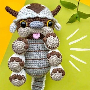 May include: A handmade crochet plush toy of a cartoon character with a brown and white head, large black eyes, and a pink tongue. The body is gray with brown stripes, and it has six brown and white paws. The toy is set against a yellow and green background.