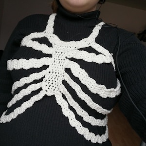 May include: A white crochet rib cage design over a black turtleneck sweater. The rib cage is made of thick, textured yarn, creating a three-dimensional effect. The design covers the chest and upper torso, offering a unique and artistic look. The sweater is a solid black color.